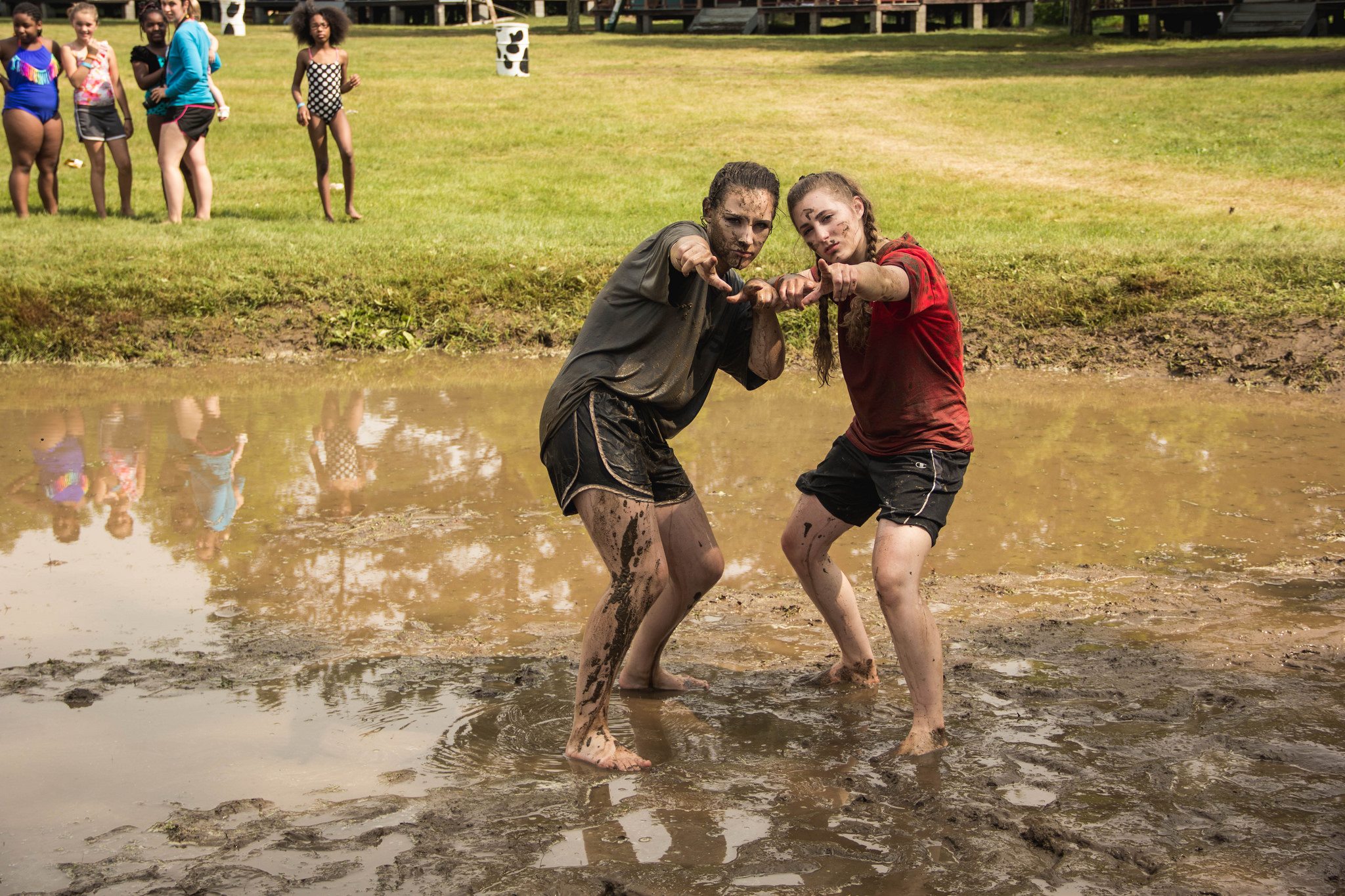 Two women standing in a mud pit pointing at the camera.