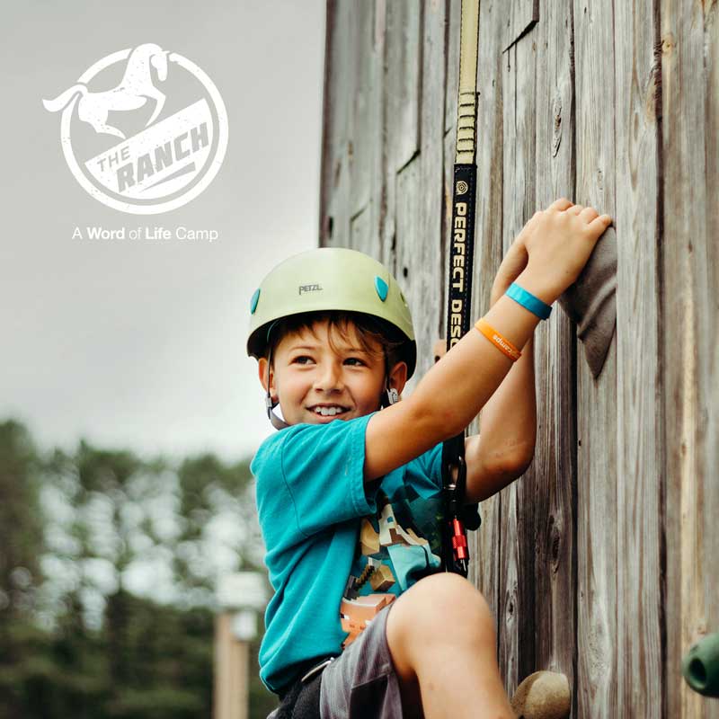 the ranch camp - kid smiling while climbing a climbing wall.