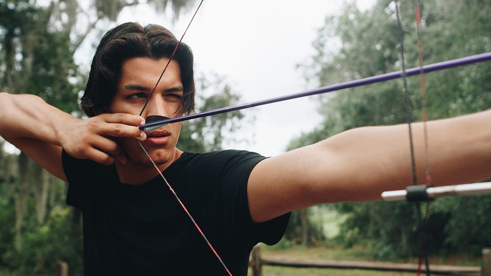 Teen looking down an arrow that he has drawn on his bow.